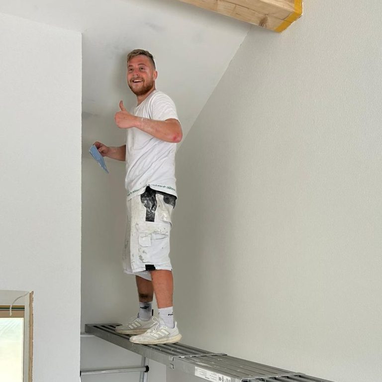 Handwerker steht auf einer Diele und streicht eine Wand mit der Geste "Daumen hoch."